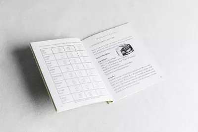 An open workbook displayed on a white surface showing a grid table on the left page and instructional text with a small image on the right page, appearing to be part of a Christmas card activity or guide from Liz Male Consulting.