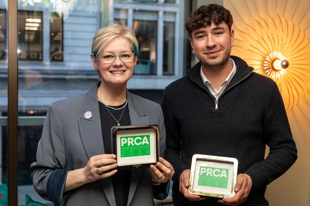 Two professionals stand together holding PRCA Workplace Champions Awards 2026 certificates, with a woman on the left wearing glasses and a grey blazer, and a man on the right in a black sweater, both smiling at the camera.