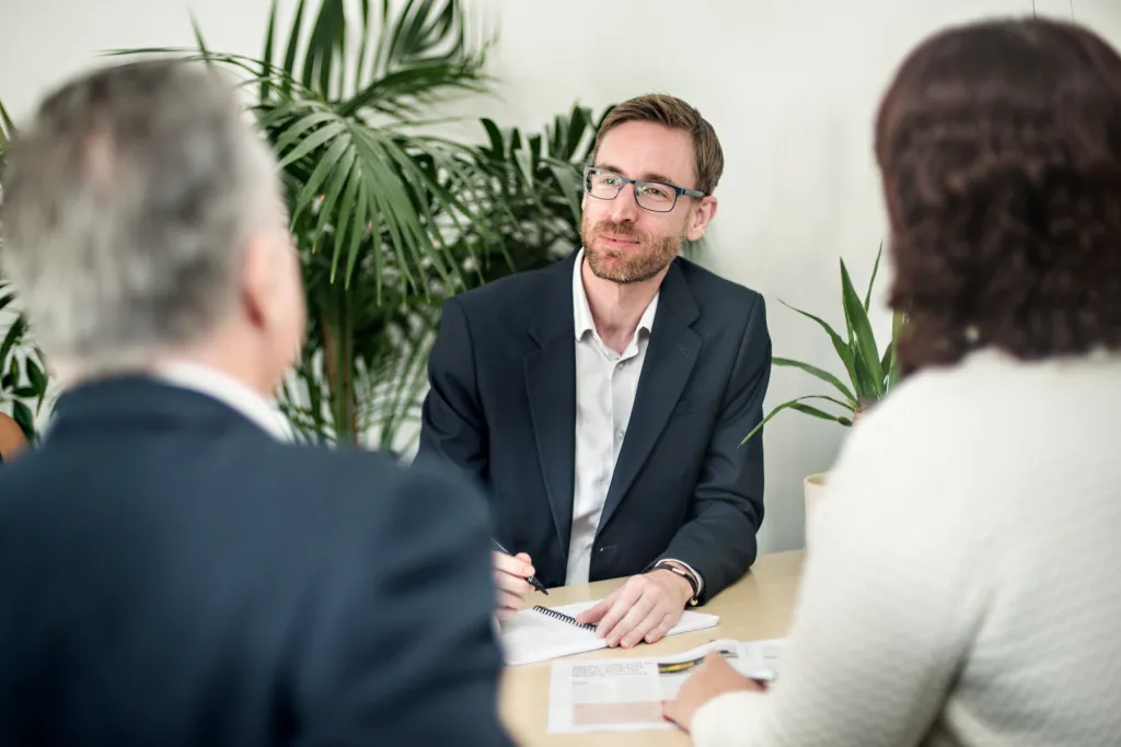 A man wearing glasses and a dark blazer sits at a table with two other people during a business meeting, with green potted plants visible in the background.