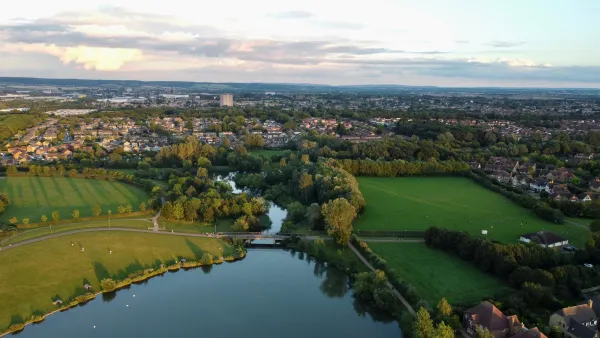 Aerial view of a river winding through green fields and residential areas with autumn foliage, suburban developments visible on both sides of the waterway under a cloudy sky.