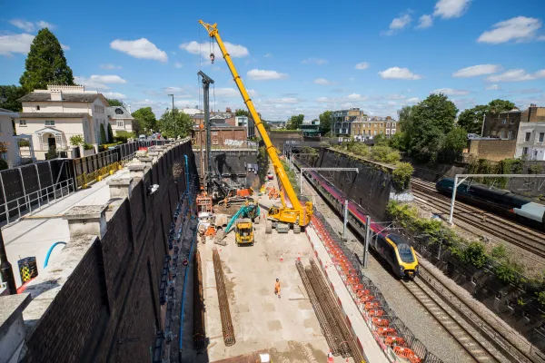 A construction site with a yellow crane positioned over an excavated pit alongside railway tracks, bordered by black safety fencing with brick buildings and trees visible in the background under a clear blue sky.
