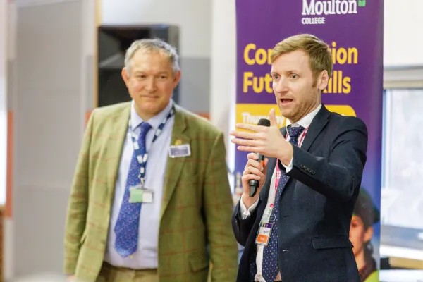 Two men stand in front of a purple Moulton College banner during a presentation. The man on the right holds a microphone and speaks to an audience, while the man on the left in a green cardigan stands beside him. Both wear conference badges and business attire.
